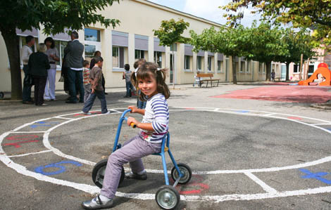 Cette photo représente une petite fille sur un tricycle. Elle est à l'arrêt et pause pour la photo. Elle se trouve dans une cours d'école. D'autres enfants et adultes sont présents en arrière plan
