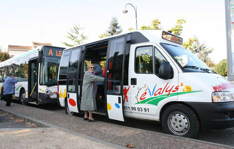 Photo représentant un arrêt de transport urbain Evalys. Une usagée âgée monte à bord d'un petit bus. D'autres usagers montent dans un bus.
