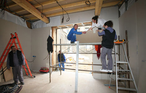 Cette photo représente des travaux d'intérieurs. On retrouve trois personnes sur une échafaudage montant une plaque. Une personne sur la gauche de la photo est près d'une échelle. Les poutres du toit sont apparentes