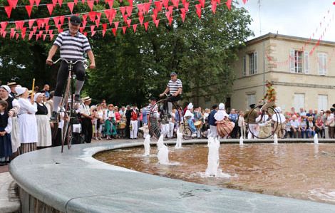 Cette photo représente un homme faisant du vélo d'époque sur les bords de la fontaine de Marmande. En arrière on retrouve dans la foule des gens déguisés en costume d'époque. Des guirlandes rouges sont accrochées dans la ville