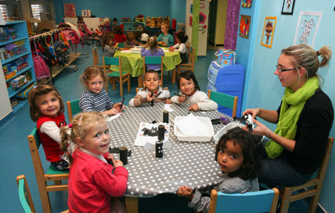 Cette photo représente des enfants assis autour d'une table ronde avec une femme. Ils font une activité. D'autres tables sont présentes dans la pièce ainsi que d'autres enfants