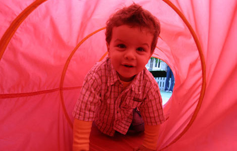 Cette photo représente un jeune enfant s'amusant en rampant dans un tuyeau en tissu rouge tendu par des cercles en plastique.