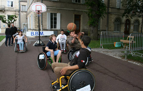Cette photo représente cinq jeunes sur fauteuil roulant jouant au basketball. Deux d'entre eux se battent le ballon.