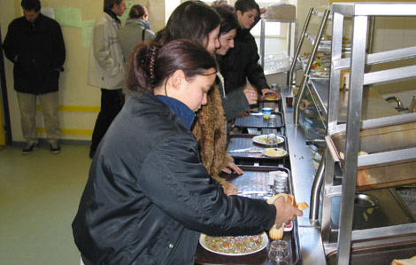 Cete photo représente un service dans un restaurant scolaire. Des jeunes font la queue pour se servir des différents plats proposés en faisant circuler leurs plateaux sur des guides en métal